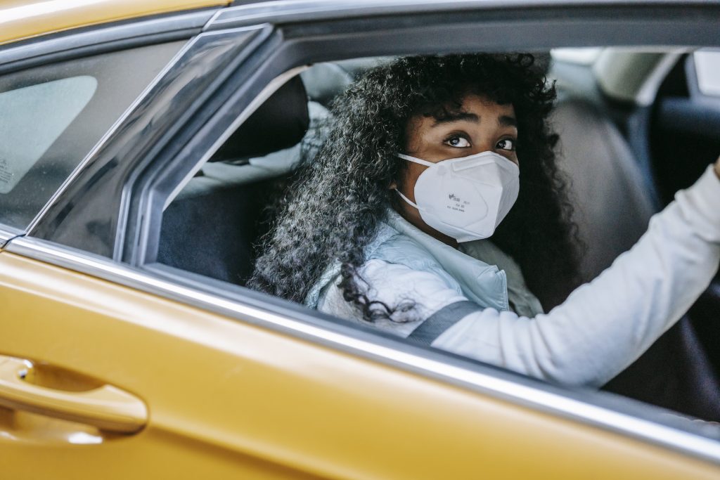 Thoughtful African American female with dark curly hair in medical protective mask sitting in taxi and looking away in daytime